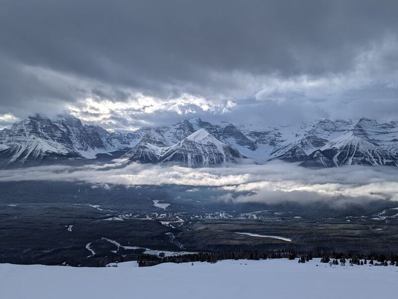 A photo from near the unload point of the Top of the World chair at the Lake Louise ski resort. The photo shows the Lake Louise village below and, between two distinct layers of cloud through which sunlight is breaking, Mount Temple, Mount Victoria, and the Victoria Glacier which feeds Lake Louise itself.